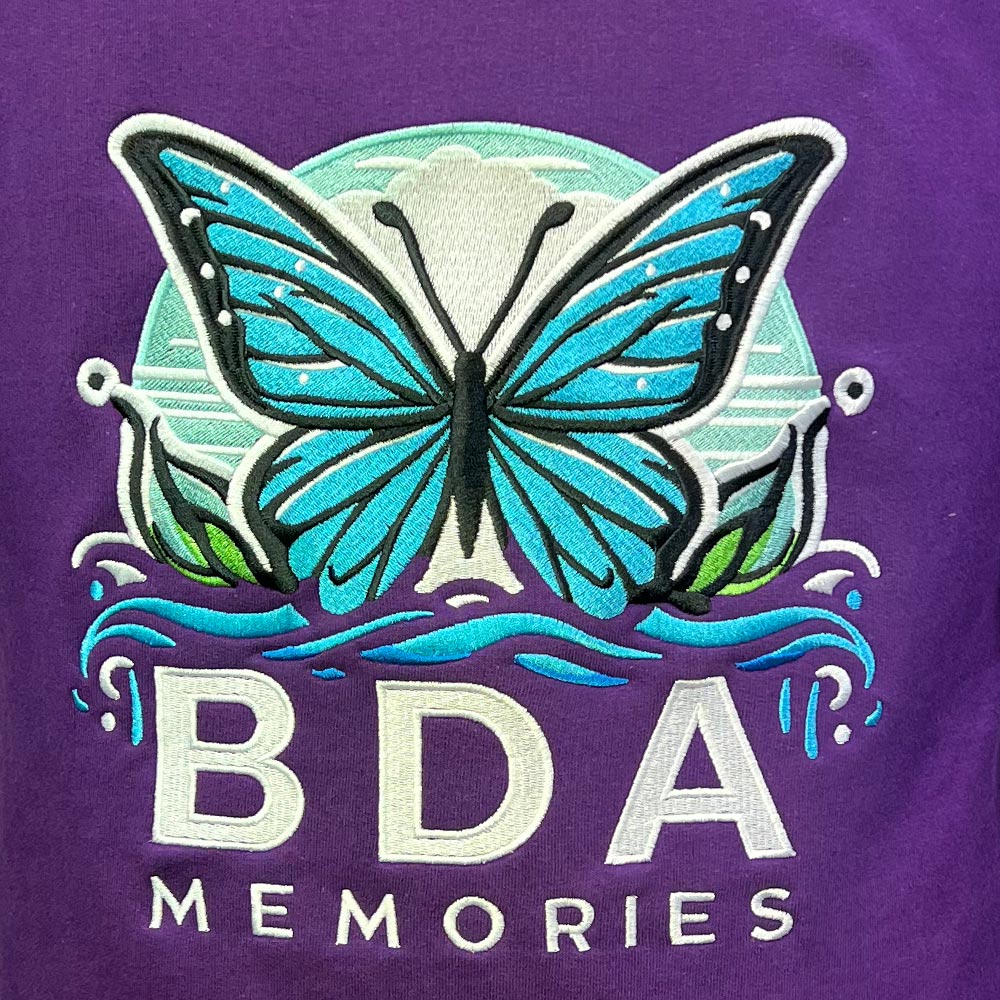 Gros plan sur la broderie papillon bleu et turquoise du logo BDA Memories réalisée sur un sweat violet.