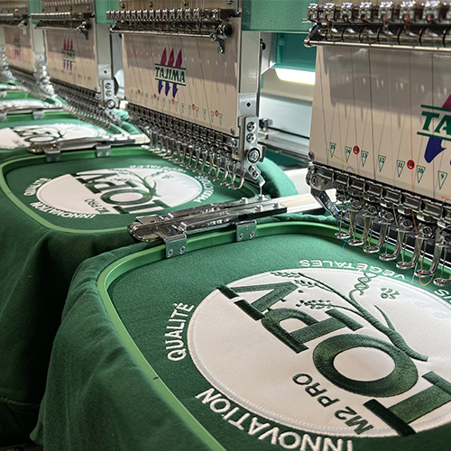 Gros plan sur une machine de broderie textile en train de marquer le logo IQPV sur des vestes sweats verts.