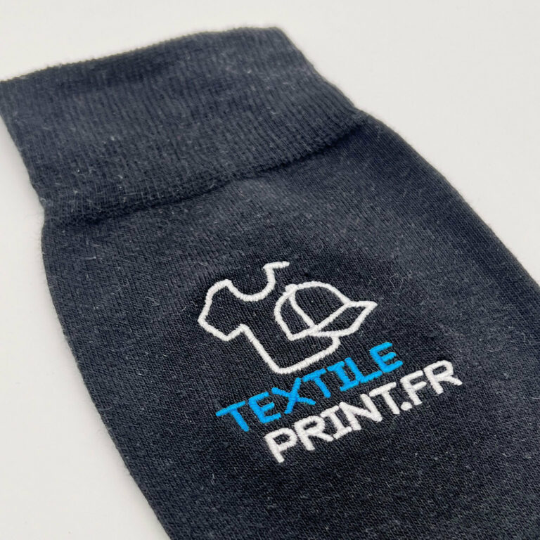 Gros plan sur le haut d'une chaussette personnalisée noire en coton peigné, arborant un logo brodé de haute qualité avec le texte "TEXTILE PRINT.FR" en bleu et blanc.