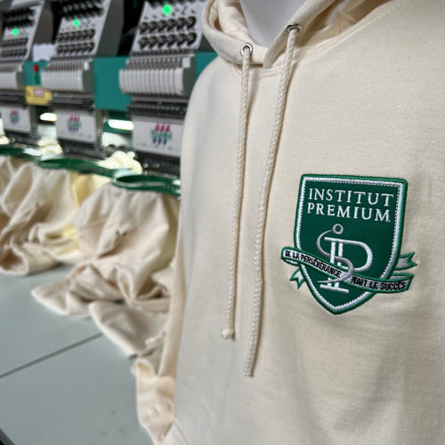 Sweat à capuche blanc écru personnalisé avec écusson brodé en position cœur, blason Institut Premium vert et blanc, prise de vue dans l'atelier